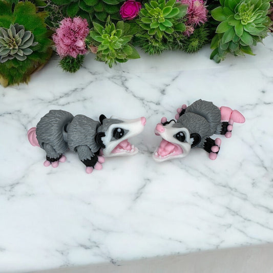 Two 3D printed baby opossum figures with gray bodies and pink accents on their faces and feet, arranged on a marble surface.
