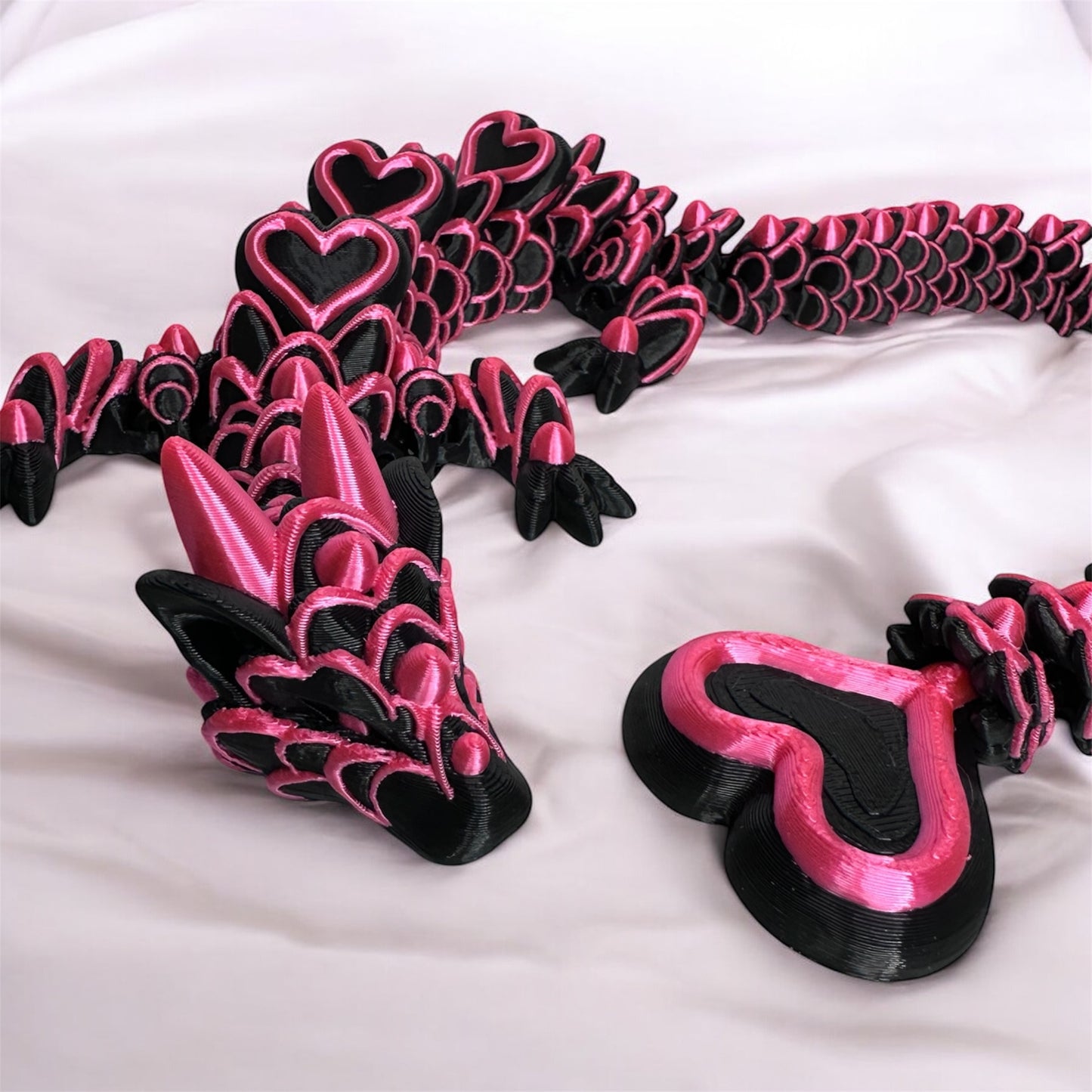 A 3d printed black articulated dragon with pink frosting and hearts down it's back. On a marble surface.