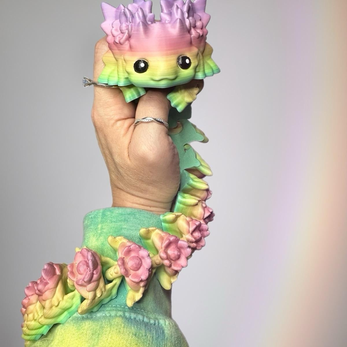 Hand holding a rainbow colored 3d printed axolotl dragon on a light colored background.