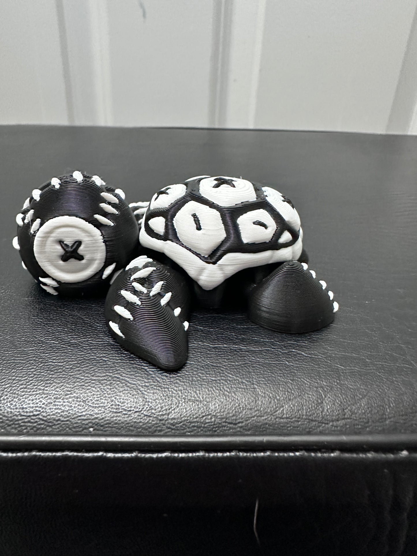 3D printed turtle that looks like its covered in in stitches. Black with white stitches. On a black surface.