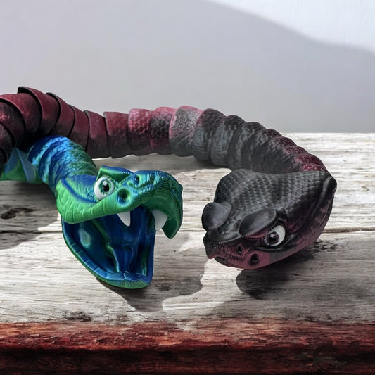 Two 3d printed rattlesnakes - one is blue/green and the other is black/red on a wood surface.