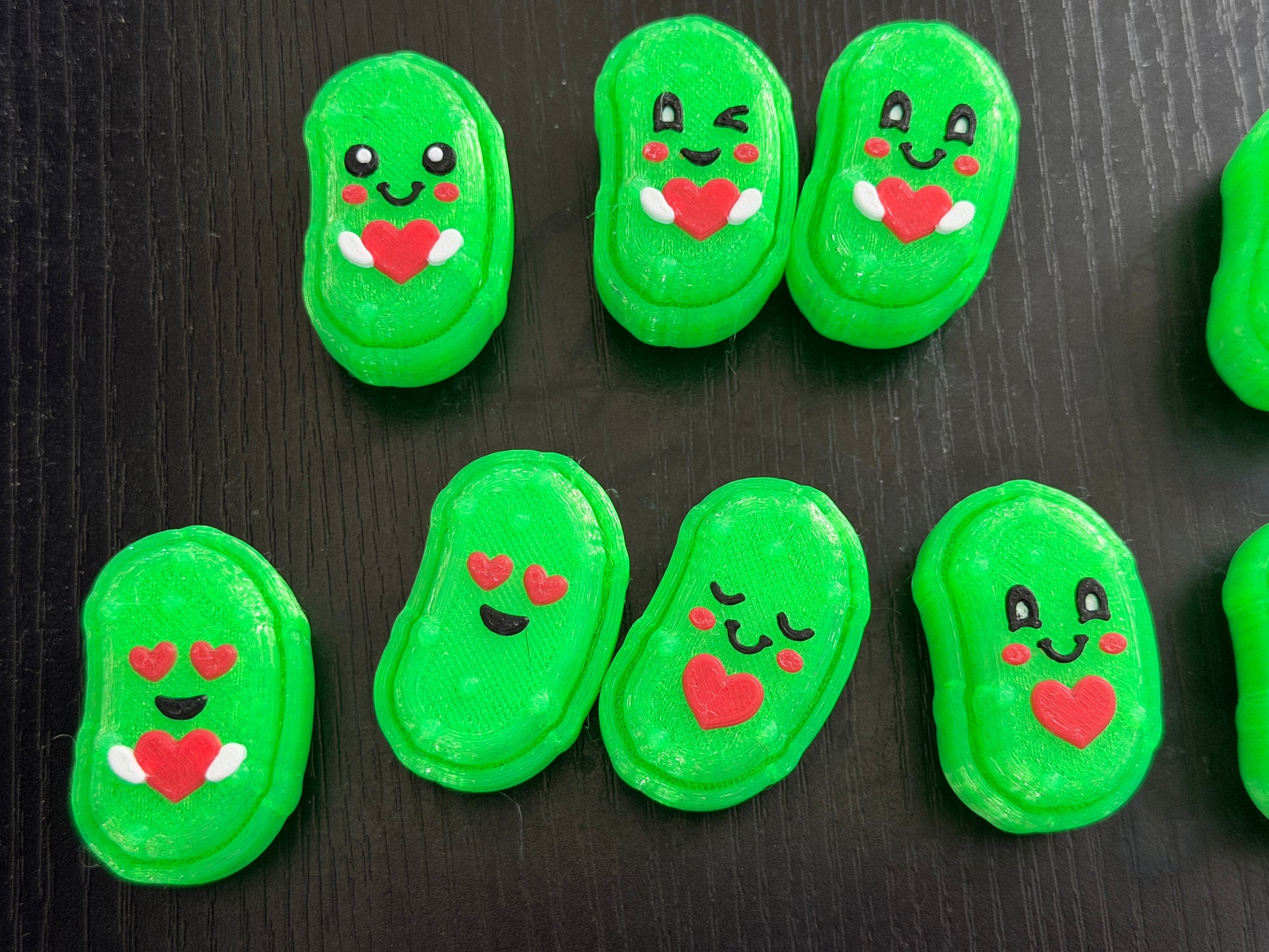 Seven 3d printed pickle shaped clicker fidgets on a black background. They all have cute faces and hearts.