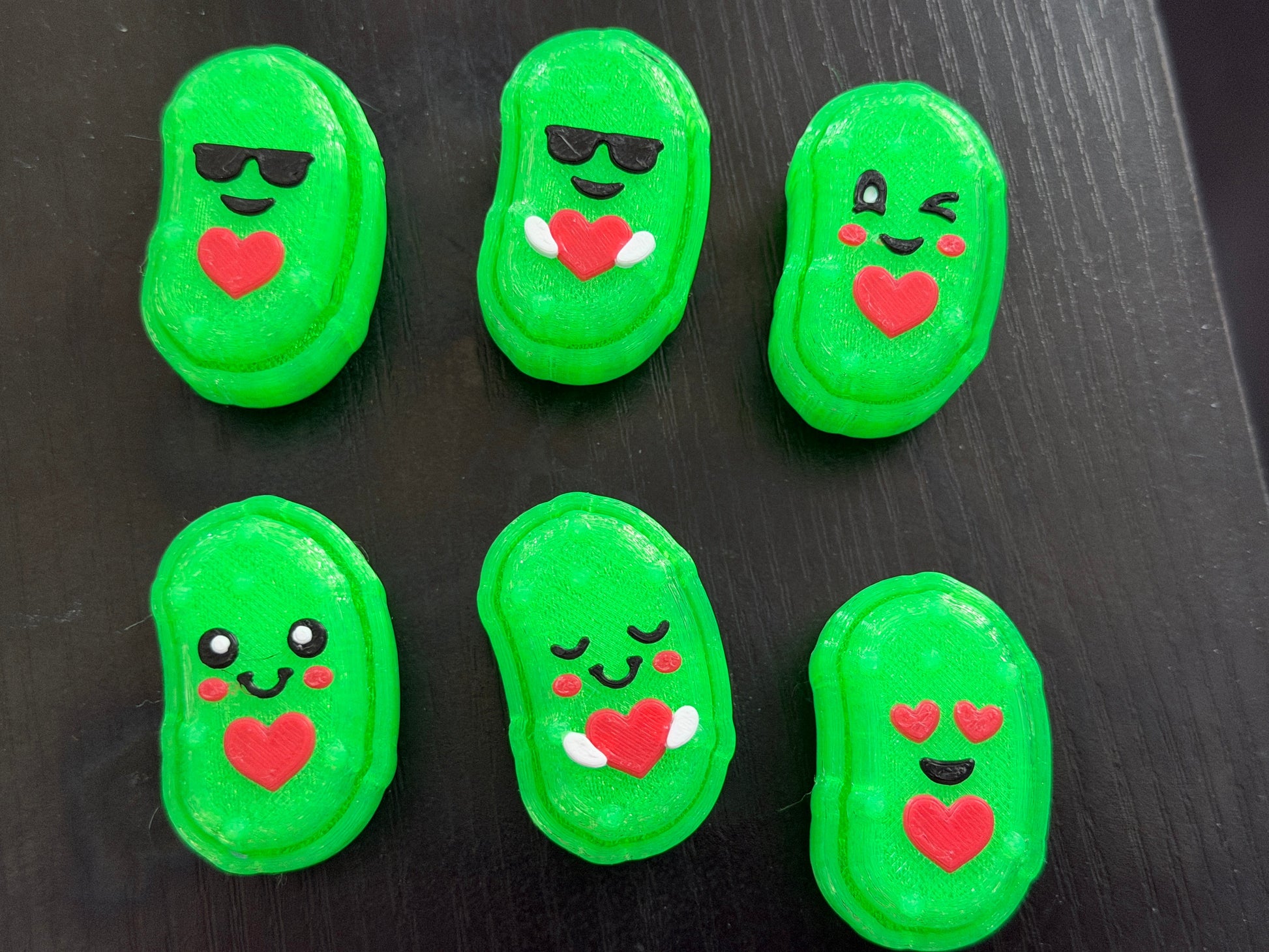 Six 3d printed pickle shaped clicker fidgets on a black background. They all have cute faces and hearts.