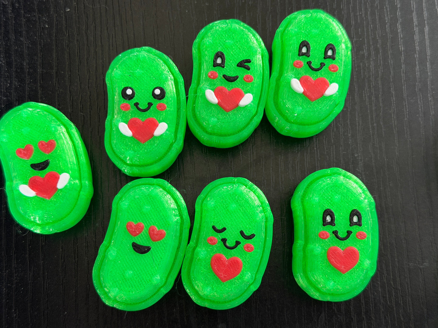 Seven 3d printed pickle shaped clicker fidgets on a black background. They all have cute faces and hearts.