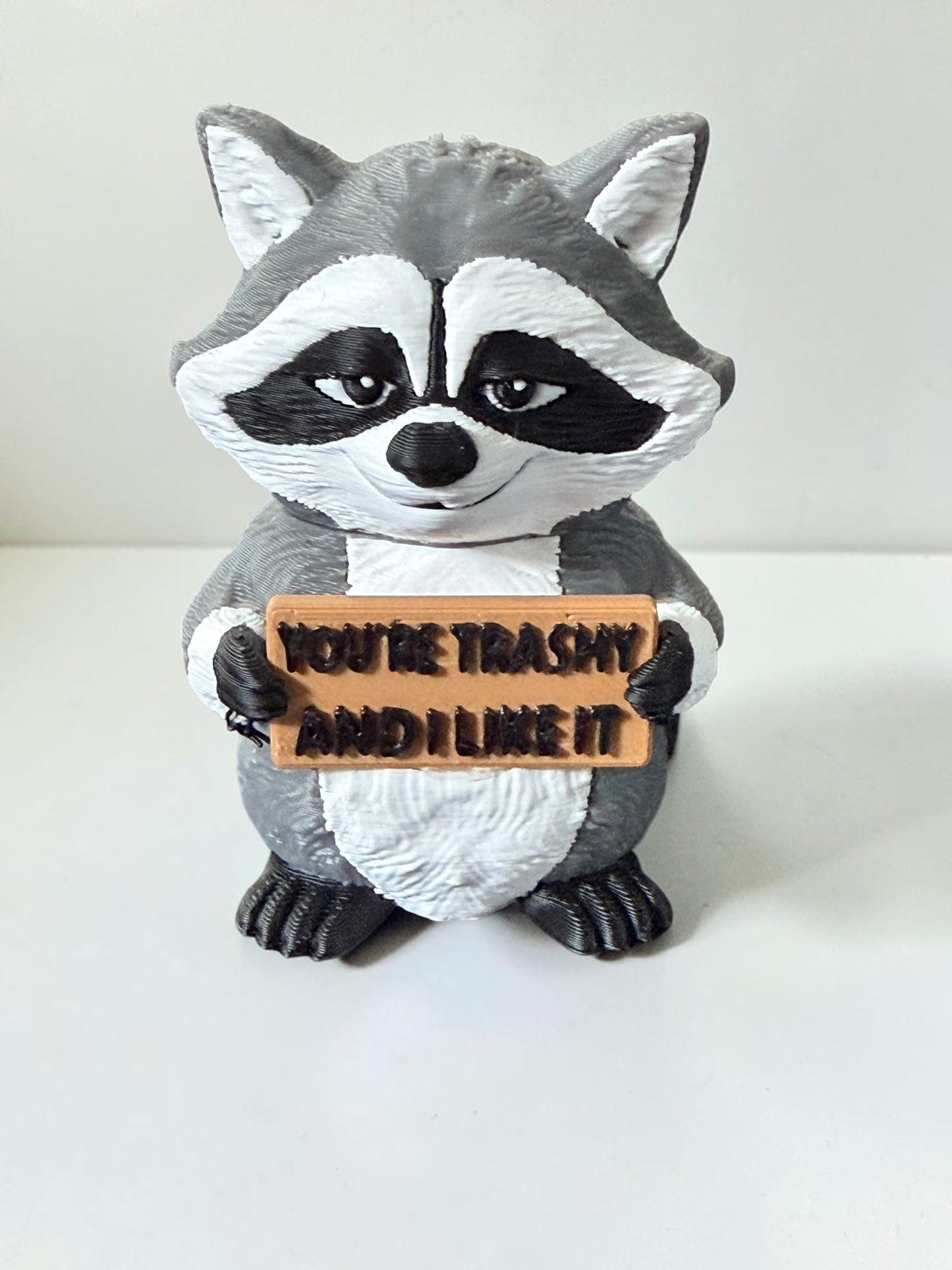 3d printed raccoon holding a sign that says "You're trashy and I like it"