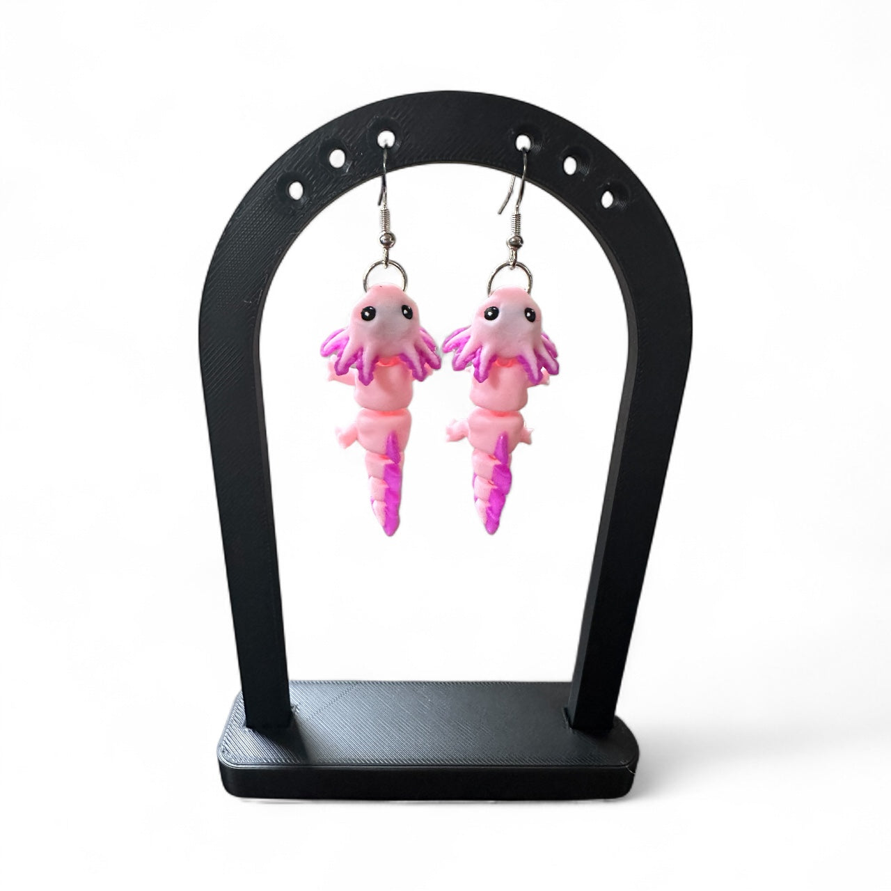 Pink axolotl-shaped earrings on a black stand against a white background
