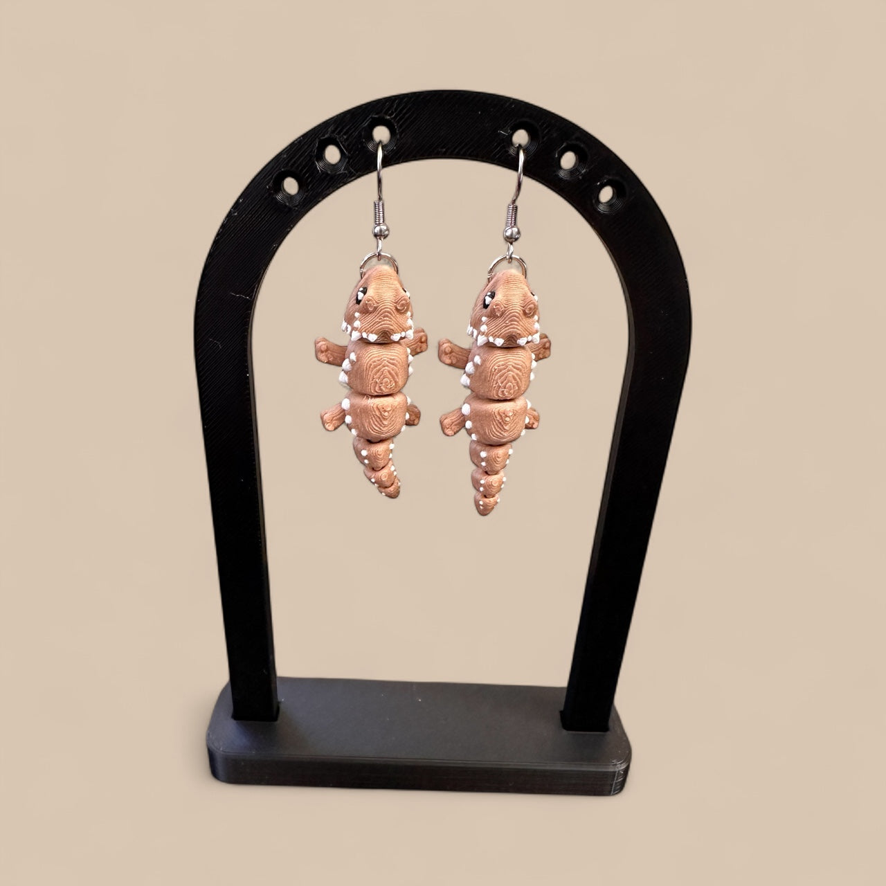 Pair of earrings shaped like bearded dragons on a black stand against a beige background