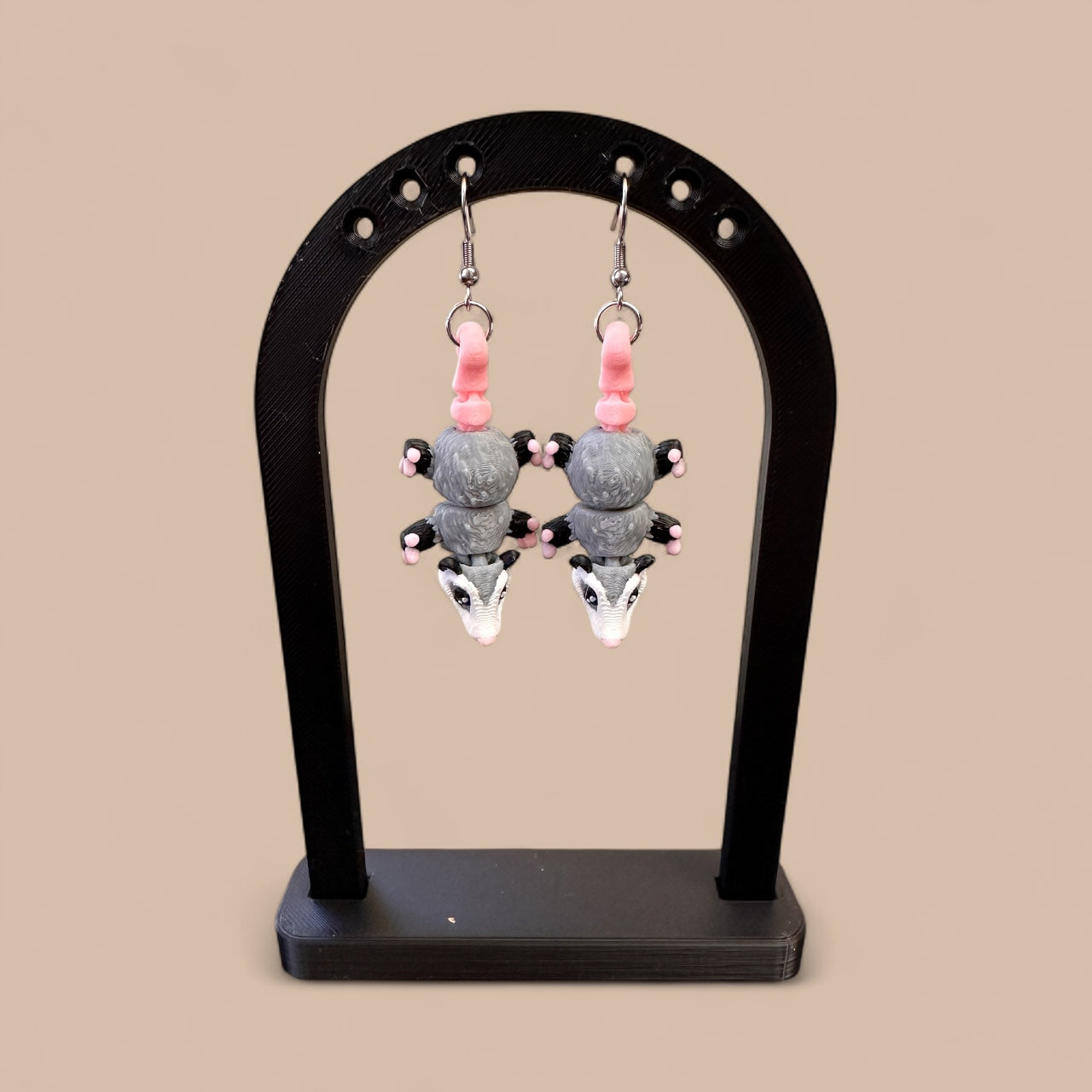 Earrings shaped like opossums on a black stand against a beige background
